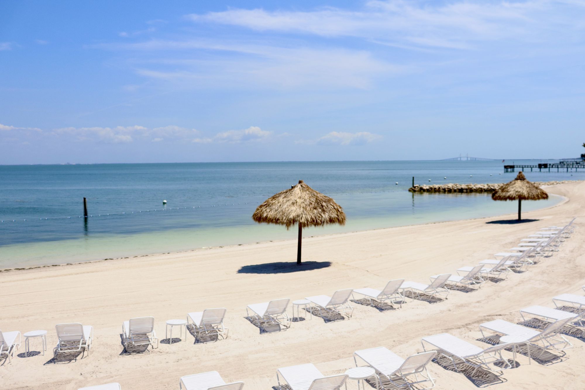 A sunny beach with white sand, lounge chairs, and thatched umbrellas beside calm blue water, with a pier extending into the sea.