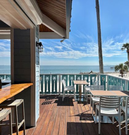 Outdoor dining area with wooden tables, chairs, and a bar counter overlooking a sunny beach and ocean, surrounded by palm trees.