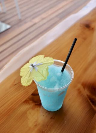 A blue slushie with a yellow paper umbrella and a black straw sits on a wooden table.