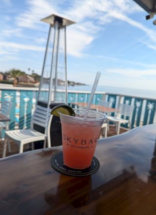 A pink drink with lime on a bar top, overlooking a scenic ocean view with chairs and a tall outdoor heater nearby.