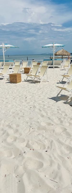 A sandy beach with white lounge chairs, small tables, and umbrellas under a cloudy blue sky, overlooking the ocean.