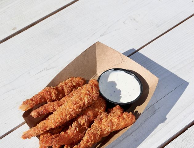 The image shows a cardboard tray with breaded chicken strips and a small cup of creamy dipping sauce on a light wooden table.