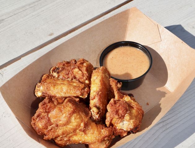 A tray of fried chicken pieces served with a small cup of dipping sauce, placed on a light-colored wooden table outdoors.