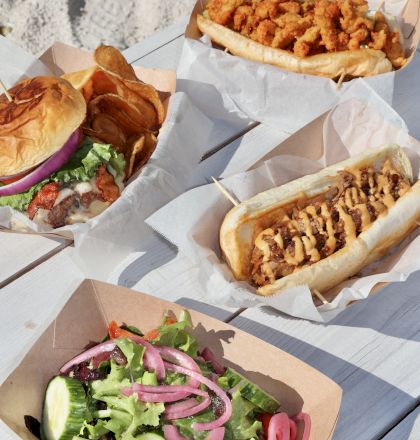 The image shows a variety of food on a table, including a burger with chips, a salad, and two types of sandwiches.