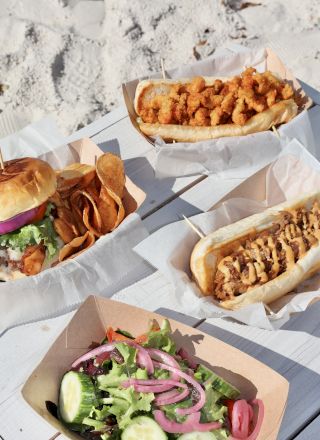 An assortment of food on a white table includes sandwiches, a salad with cucumbers, and a side of chips or fries, set on a sandy beach.