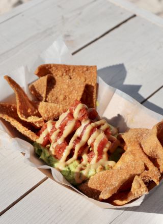 A paper tray with tortilla chips and a topping drizzled with sauce, placed on a white wooden table, in a sunny outdoor setting.