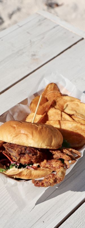 A crispy chicken sandwich with lettuce in a bun is served with potato chips on a rectangular tray placed on a white wooden table outdoors.