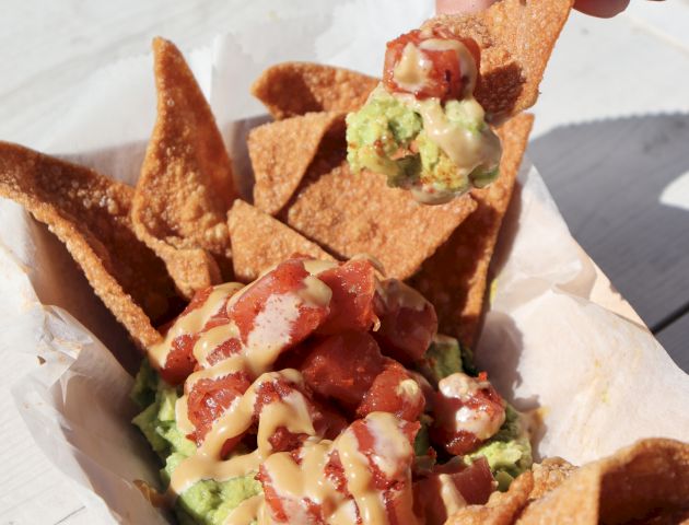 A hand reaches for a tortilla chip topped with avocado and salsa. The chips are in a basket on a white wooden surface.