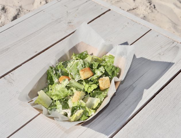 A Caesar salad with lettuce, croutons, and dressing is served in a paper dish on a wooden table near a sandy area.