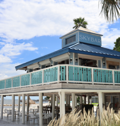 A beachside bar with a turquoise railing and sign reading "SkyBar," set against a backdrop of palm trees and a partly cloudy sky.