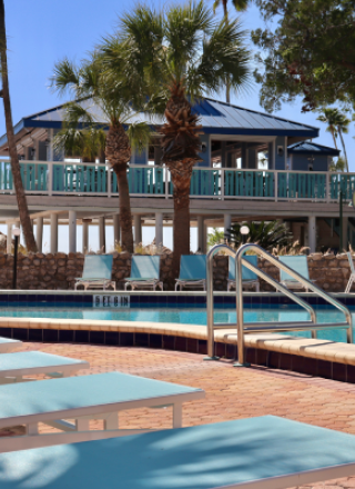 Lounge chairs and a pool with umbrellas are in front of a blue-roofed building surrounded by palm trees.