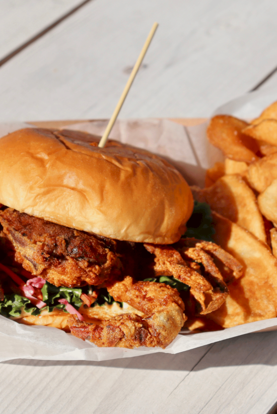 A sandwich with a soft-shell crab, toppings, and a skewer, accompanied by a side of thick-cut potato chips on a wooden table.