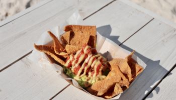 Crispy tortilla chips served with a topping of guacamole and drizzled sauces, placed on a paper tray on a white wooden table.