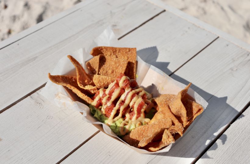 Crispy tortilla chips served with a topping of guacamole and drizzled sauces, placed on a paper tray on a white wooden table.