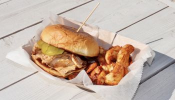 A sandwich with pickles and grilled meat, served alongside seasoned shrimp, sits on a wooden table in a paper basket.