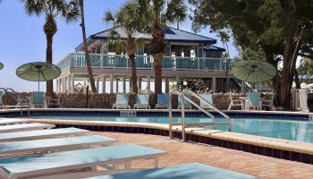 A serene poolside scene with lounge chairs, umbrellas, and a blue-roofed building surrounded by palm trees in the background.